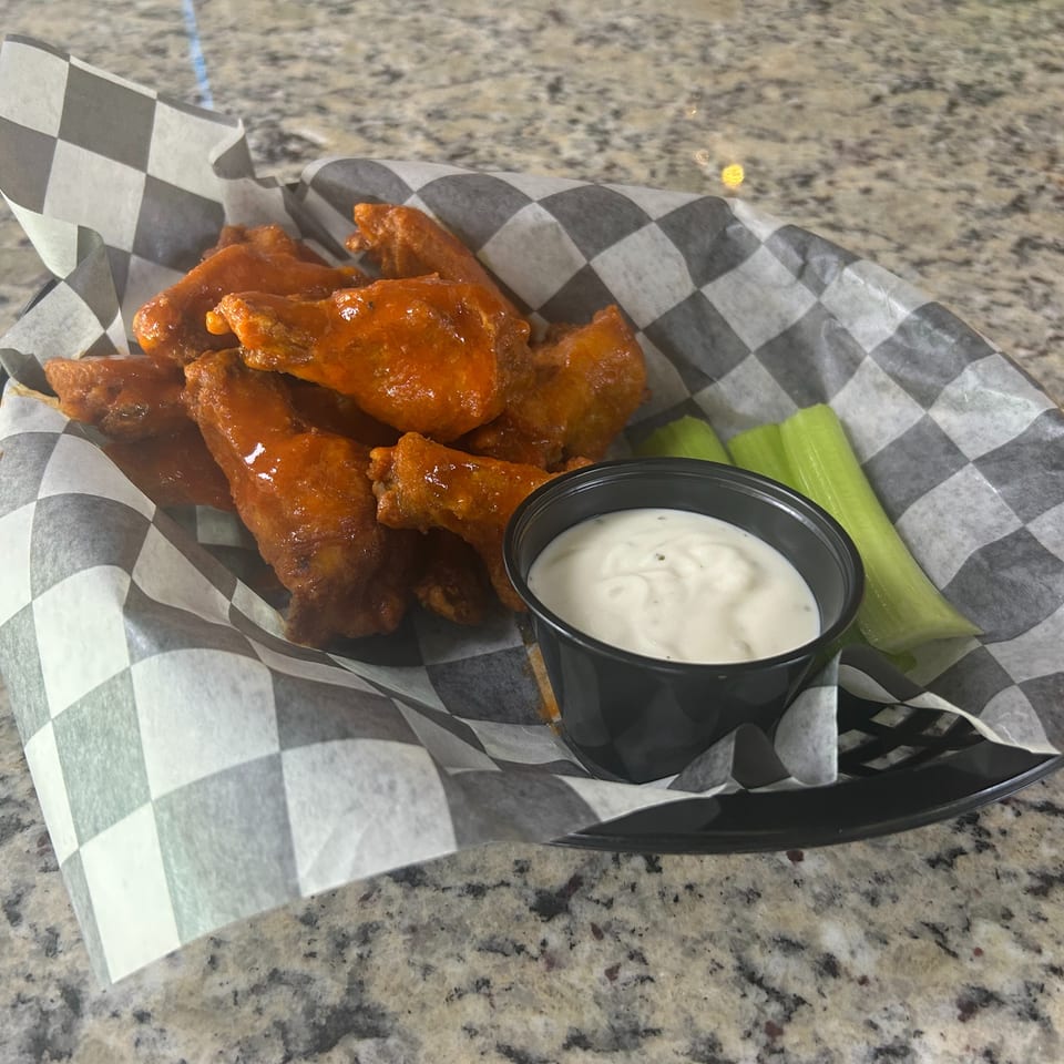 Buffalo chicken wings served with celery sticks and creamy dipping sauce in a checkered paper-lined basket.